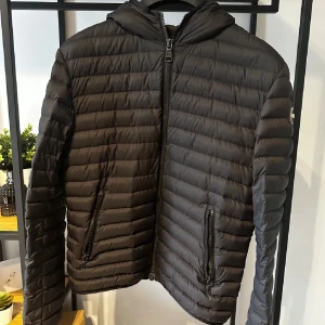 Svart colmar jacka - Snygg svart pufferjacka med huva och dragkedja framtill. Jackan har långa ärmar och två praktiska fickor med dragkedja. Perfekt för kyligare dagar.