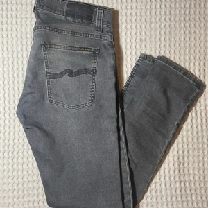 Grå jeans från Nudie Jeans - Snygga grå jeans från Nudie Jeans med klassisk femficksdesign och dragkedja. De har en broderad detalj på bakfickan och är tillverkade i denim av hög kvalitet. Perfekta för en stilren look. Kom me bud