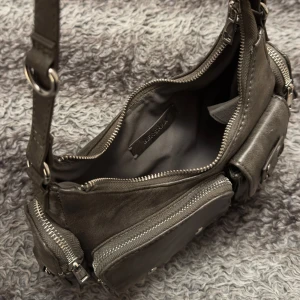 Vintage väska från Bershka - Snygg grå axelväska i syntetmaterial med flera praktiska fickor och dragkedjor. Väskan har en justerbar axelrem och dekorativa metalldetaljer. Perfekt för att hålla ordning på dina saker med stil.