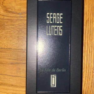 La Fille de Berlin Parfym från Serge Lutens - Elegant parfymflaska från Serge Lutens med namnet 'La Fille de Berlin'. Flaskan är rektangulär med en djup röd vätska och en svart etikett. Korken är svart och avtagbar med en sprayfunktion. Perfekt för den som söker en sofistikerad doftupplevelse.
