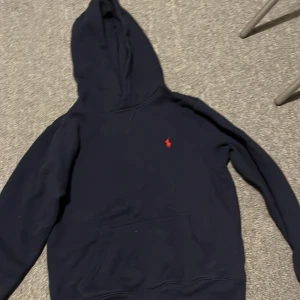 Mörkblå hoodie från Ralph Lauren 10-12 - Säljer en stilren mörkblå hoodie från Ralph Lauren med det klassiska röda logotypbroderiet på bröstet. Tröjan har en bekväm passform med långa ärmar och en praktisk magficka. Perfekt för en avslappnad look. Priset är inte spikat i sten 