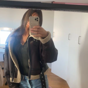 Svart/brun skinnjacka med pälsdetaljer - Snygg svart/brun skinnjacka med mysiga pälsdetaljer i beige. Jackan har dragkedja och bälte i midjan för en cool look. Perfekt för kyligare dagar.