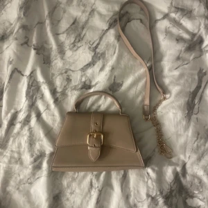 Beige handväska med axelrem - Snygg beige handväska med justerbar axelrem och dekorativt spänne framtill. Skriv om man har frågor ❤️