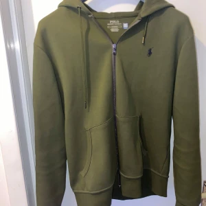 Olivgrön hoodie från Polo Ralph Lauren - Säljer en stilren olivgrön hoodie från Polo Ralph Lauren. Den har en dragkedja framtill och en klassisk logga på bröstet. Perfekt för en avslappnad look med sina långa ärmar och bekväma passform.