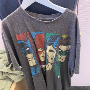 Grå t-shirt med DC superhjältar - Säljer en grå t-shirt med tryck av tre kända superhjältar i färgerna blå, röd och grön. T-shirten har en normal passform och korta ärmar. Perfekt för alla som älskar serietidningar och superhjältar!