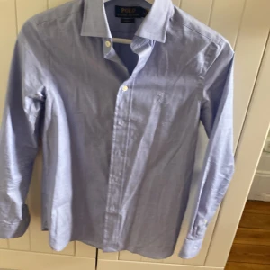 Blå skjorta från Ralph Lauren - Säljer en stilren blå skjorta från Ralph Lauren i slim fit. Skjortan har långa ärmar och knappar framtill samt vid ärmsluten. Perfekt för en elegant look.