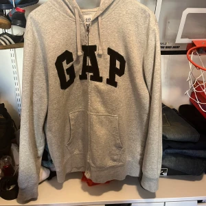 Grå hoodie från GAP - Säljer en klassisk grå hoodie från GAP med dragkedja och stor logga på bröstet. Tröjan har en bekväm passform med långa ärmar och en praktisk huva med dragsko. Perfekt för en avslappnad stil.