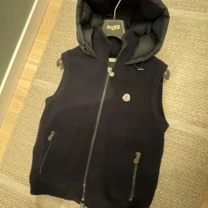 Marin blå stickad väst med huva från Moncler - Snygg mörk blå stickad väst från Moncler med en snygg huva. Västen har dragkedja framtill och två fickor med dragkedjor. Perfekt för lager-på-lager under jumna dagar som en fin vår, höst dag. Västen är i storlek M (passar S) och passar till allting. Skriv vid funderingar!