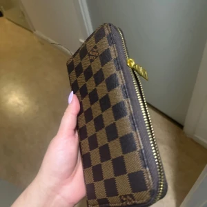 Rutig plånbok med dragkedja - Snygg plånbok med klassiskt rutmönster i brunt och beige. Den har en praktisk dragkedja och flera fack för kort och kontanter. Perfekt för att hålla ordning på dina tillhörigheter med stil. 1 för 120kr 2 för 200kr.