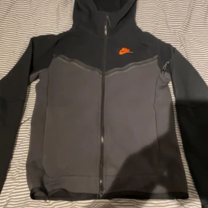 Svart och grå Nike hoodie  - Säljer en svart och grå huvtröja från Nike med orange logga på bröstet. Tröjan har dragkedja och  Passar dig som gillar sportig design. Den är i väldigt bra skick och kostar egentligen 1500 kr. Men mitt pris är bara 600 kr.