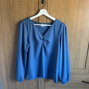 Blå v-ringad blus - Snygg blå blus med v-ringning och långa ärmar. Blusen har en  rynkning framtill. Perfekt för både vardag och festliga tillfällen. Från Vila 