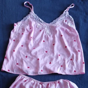 Rosa pyjamasset med hjärtan från H&M - Säljer ett sött rosa pyjamasset från H&M med små röda hjärtan och spetsdetaljer. Linnetoppen har smala axelband och spets vid halsringningen. Shortsen har elastisk midja och spets vid bensluten. Perfekt för mysiga hemmakvällar! 💖