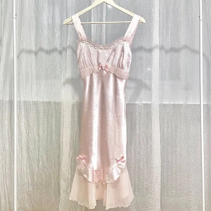 Ljusrosa satinklänning med volanger och rosetter - Charmig ljusrosa klänning i siden med hjärtmönster. Den har volanger och söta rosetter i midjan och vid fållen. Blommiga spetsdetaljer pryder halsringningen. Ge en fairy-look som passar väldigt bra till våren och sommaren.