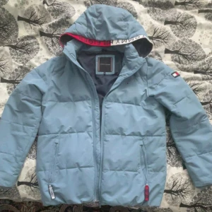 Ljusblå dunjacka från Tommy Hilfiger - Snygg ljusblå dunjacka från Tommy Hilfiger med huva och dragkedja. Jackan har märkets logga på ärmen och insidan av huvan. Perfekt för kyliga dagar!