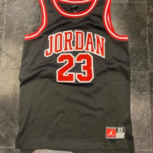 Svart basketlinne med Jordan 23 - Snyggt svart basketlinne med röd och vit text 'Jordan 23' på framsidan. Linnet har röda och vita detaljer runt ärmhålen och halsen. Perfekt för basketentusiaster eller som en cool streetwear-look.