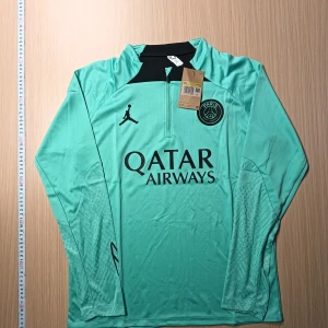 Grön långärmad fotbollströja från Jordan - Snygg grön långärmad fotbollströja från Jordan med svart logga och text. Perfekt för träning eller match. Tröjan har en dragkedja vid halsen och klubbmärke på bröstet.