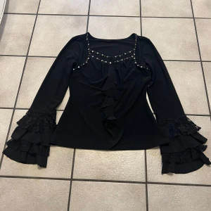 Svart blus med volang och spetsdetaljer storlek S/M - Elegant svart blus med långa ärmar och volangdetaljer framtill. Blusen har spetsdetaljer på ärmsluten och dekorativa knappar längs halsringningen. Perfekt för en stilfull look.
