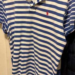 Blåvit randig pikétröja från Ralph Lauren - Snygg blåvit randig pikétröja från Ralph Lauren med klassisk krage och knappdetalj. Perfekt för en avslappnad och stilren look. Den har korta ärmar och en liten röd logotyp på bröstet.