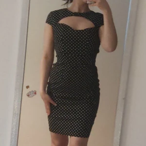 Svart prickig klänning från Hell Bunny Vixen - Snygg svart klänning med vita prickar från Hell Bunny. Klänningen har en unik cut-out detalj vid halsen och korta ärmar. Perfekt för en retro look!