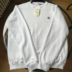 Vit sweatshirt från Moncler - Säljer en stilren vit sweatshirt från Moncler med rund halsringning och diskret Moncler-logga broderad på bröstet. Tröjan har ribbade muddar vid ärmslut och nederkant. Perfekt för en clean och klassisk look. Den är aldrig använd!