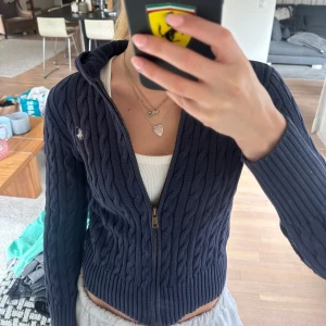 Mörkblå kabelstickad hoodie från Ralph Lauren - Säljer en mörkblå stickad hoodie från Ralph Lauren med dragkedja framtill och kabelstickat mönster. Tröjan har långa ärmar, huva och den klassiska loggan broderad på bröstet. 