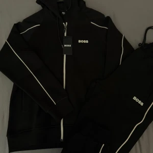 Svart hoodie från BOSS med vita detaljer - Säljer en svart hoodie från BOSS med dragkedja och vita ränder längs ärmarna. Tröjan har huva med snörning och BOSS-logga på bröstet. Perfekt för en sportig och stilren look. Storlek S M L är tyvärr sålda men vi har just nu i XL XXL