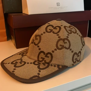 Beige keps från Gucci med GG-mönster - Säljer en beige keps från Gucci med det klassiska GG-mönstret i brunt. Kepsen har justerbar rem baktill och är tillverkad i bomullsmaterial. Perfekt för dig som vill ha en snygg och trendig accessoar med lyxig känsla.