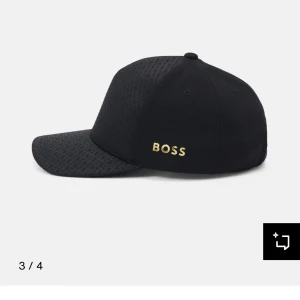 Svart keps från BOSS - Stilren svart keps från BOSS med diskret mönster på skärmen och guldigt BOSS-tryck på sidan. Klassisk form med böjd skärm och justerbar passform. Perfekt accessoar för en sportig och modern look.