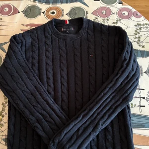 Mörkblå tröja från Tommy Hilfiger (164) - Säljer en mörkblå kabelstickad tröja från Tommy Hilfiger. Tröjan har rund halsringning, lång ärm och diskret logga på bröstet. Klassisk och stilren modell som passar perfekt till kyligare dagar.
