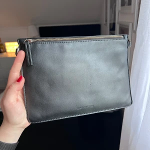 Svart väska från Carin Wester - Stilren svart väska från Carin Wester. Väskan har silverfärgad dragkedja upptill och flera fack inuti för enkel organisering. Perfekt för dig som gillar minimalistisk design och vill ha en praktisk accessoar. Finns ett litet märke på framsidan (syns på första bilden). Måtten är 18,5x26cm ❤️ (Ps. jag säljer även två stycken olika axelremmar separat som går att köpa till. ☺️)