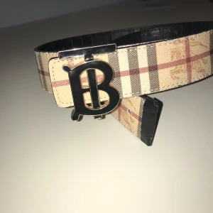 Burberry bälte med monogramspänne - Snyggt Burberry bälte med stort silverfärgat monogramspänne. Ena sidan har klassiskt beige rutigt mönster och den andra är svart med diskret rutmönster. Perfekt accessoar för att lyfta din outfit.