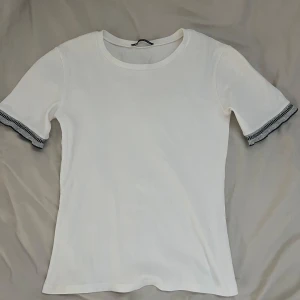 Vit topp med detaljer från Zara - Säljer en vit topp från Zara med korta ärmar och detaljer i svart och grått vid ärmsluten. Tröjan är i bra skick och säljer pga att den aldrig kommer till användning.