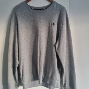 Grå sweatshirt från U.S. Polo Assn. - Klassisk grå sweatshirt från U.S. Polo Assn. med rund halsringning och broderad logga på bröstet. Tröjan har långa ärmar och ribbade muddar vid ärmslut och nederkant. Perfekt för en avslappnad stil.