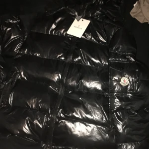 Svart pufferjacka från Moncler (äkta) - Säljer en glansig svart pufferjacka från Moncler med huva och dragkedja. Jackan har alla etiketter och lappar kvar och Moncler-logga på ärmen och olivgrön insida. Perfekt för kalla dagar när du vill hålla stilen.