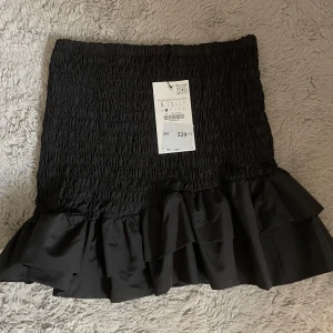 Svart volangkjol - Supersnygg svart kjol med smock upptill och dubbla volanger nertill. Kjolen är kort och har en trendig, feminin look som passar perfekt till en cool outfit. Materialet känns mjukt och har en lätt glansig finish.