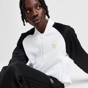 Vit och svart track jacket/tröja från Adidas - Säljer en stilren track jacket från Adidas med vit front och rygg samt svarta ärmar. Den har dragkedja och guldigt Adidas-tryck på bröstet. Helt ny och oanvänd med prislapp på den. Fick den i present fast storleken var lite fel så försöker sälja den.