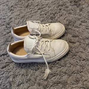 Vita sneakers med dragkedja från guiseppe zanotti - Säljer ett par vita sneakers med snörning och dragkedja på sidan. Skorna har rund tå och platt sula, vilket gör dem både snygga och bekväma. Perfekta till vardags för en clean och stilren look.