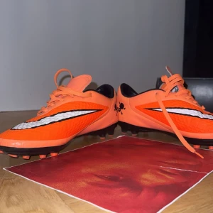 Nike Hypervenom orange fotbollsskor - Säljer ett par orangea Nike Hypervenom fotbollsskor med svart och vit swoosh på sidan. Skorna har snörning, låg profil och mönstrad ovandel för bättre bollkontroll. Svart dödskalle-detalj på hälen och svarta sulor med dobbar.