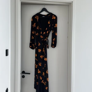 Svart omlottklänning med orange blommor från Ganni - Säljer en svart lång omlottklänning från Ganni med orange blommigt mönster. Klänningen har långa ärmar, v-ringning och knyt i midjan. Stl 38 men jag är en 36:a och tycker den passar mig bra. 