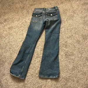 Blå bootcut jeans från Gina Tricot - Säljer ett par klassiska blå jeans från Gina Tricot i bootcut-modell. Jeansen har normal passform, dragkedja och dubbla knappar framtill samt fickor med lock och knapp bak. Perfekta för dig som gillar en tidlös jeansstil.
