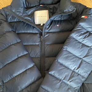 BLÅ U ESSENTIAL LIGHT DOWN JACKET - Säljer en mörkblå pufferjacka från Tommy Hilfiger. Jackan har dragkedja framtill. Perfekt och enkel att matcha till olika outfits. Säljer den då jackan blivit för liten. Den har ett litet hål på armen som man kan se på sista bilden. Pris går att diskutera