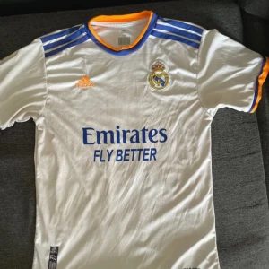 Real Madrid hemmatröja 2021/22 från Adidas - Vit Real Madrid fotbollströja med blå och orange detaljer, Adidas-logga och klubbmärke på bröstet. Kortärmad modell med blå ränder på axlarna och sponsortryck på framsidan. Tillverkad i lätt och ventilerande material.