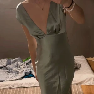 Ljusgrön långklänning med djup v-ringning - Säljer en elegant ljusgrön långklänning med djup v-ringning och öppen rygg. Klänningen har en omlottliknande detalj bak och är tillverkad i ett glansigt, mjukt material som ger en lyxig känsla. Perfekt för bal eller festliga tillfällen.