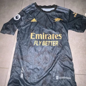 Arsenal bortatröja Ødegaard 8 - Snygg svart Arsenal bortatröja med guldiga detaljer från Adidas. Tröjan har korta ärmar, klubbmärke och sponsortryck på bröstet samt Ødegaard och nummer 8 på ryggen. Tillverkad i lätt och ventilerande material.