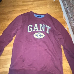 Vinröd sweatshirt från GANT - Säljer en vinröd sweatshirt från GANT med broderad logga på bröstet i vitt och gult. Tröjan har rund halsringning och långa ärmar. Perfekt för en avslappnad och stilren look.