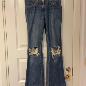 Low waist bootcut jeans med hål - Säljer ett par blå bootcut jeans med hål på båda knäna. Klassisk femficksmodell med normal passform och låg midja. Sjukt snygga men tyvärr för små för mig!