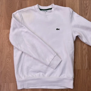 Vit sweatshirt från Lacoste - Säljer en vit sweatshirt från Lacoste med ikoniska krokodilloggan. Tröjan har långa ärmar och är som ny. Den är varm, super bra kvalite och snygg.