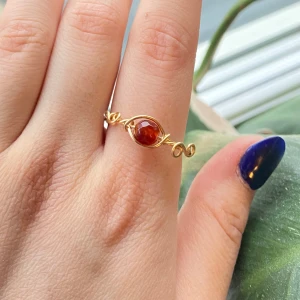 Handgjord guldpläterad ring med röd sten - Unik ring i guldpläterad metall med en vackert infattad rund röd sten i mitten. Ringen har en elegant, snirklig design som ger ett bohemiskt och trendigt intryck. Perfekt accessoar för att lyfta din stil.