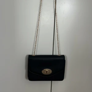 Svart axelväska med guldkedja - Snygg svart axelväska med guldfärgad kedja och ett elegant vridlås i guld på framsidan. Väskan har en stilren och klassisk design som passar perfekt till många olika outfits.