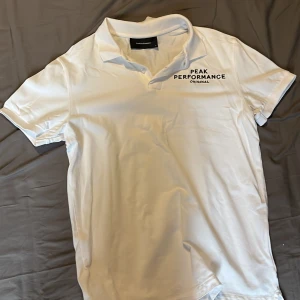 Vit piké från Peak Performance - Säljer en klassisk vit pikétröja från Peak Performance med svart logga på bröstet. Tröjan har krage och korta ärmar, perfekt för en stilren och sportig look.